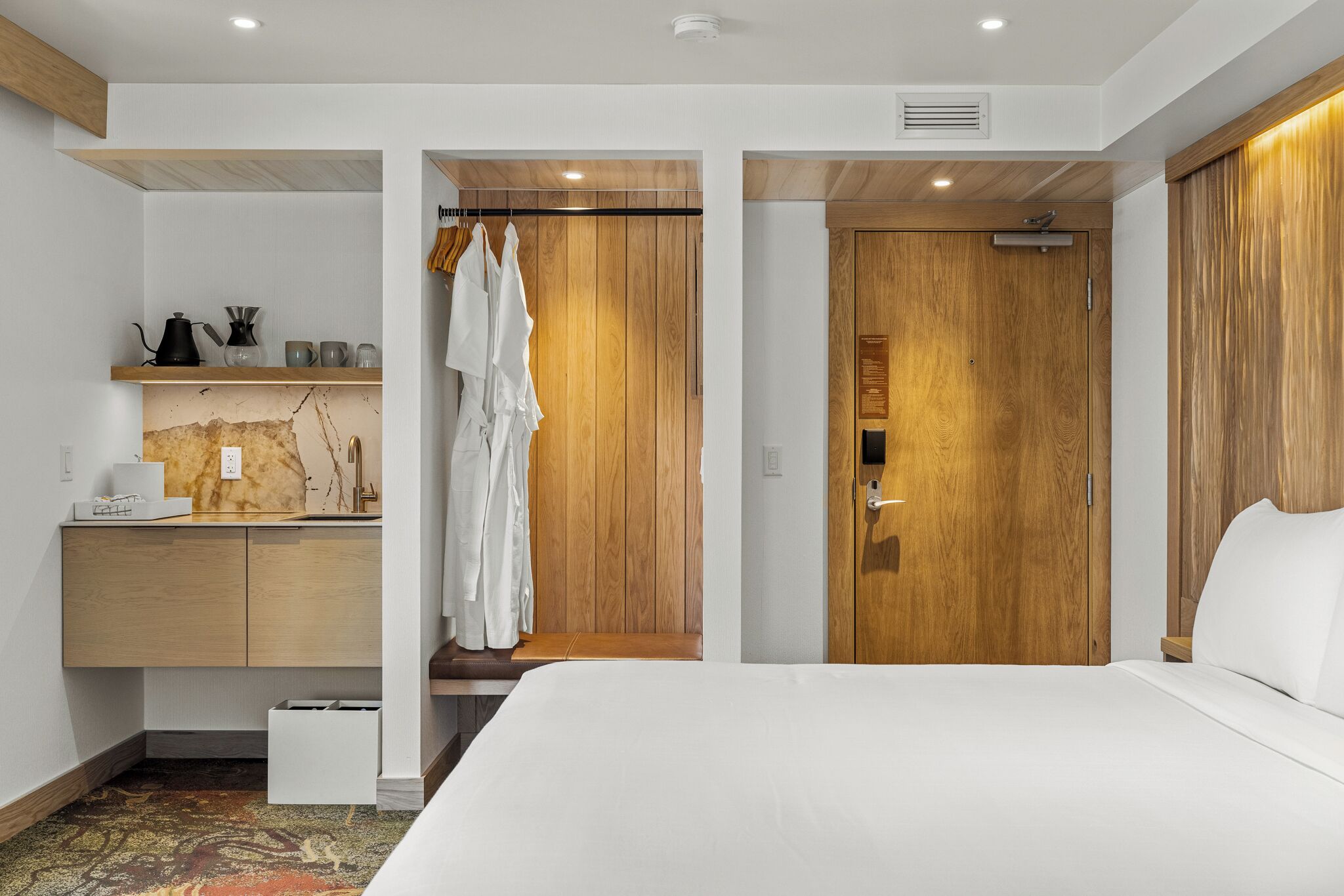 A modern hotel room with a wooden wardrobe, a bed, a small desk with a kettle, and a bench.