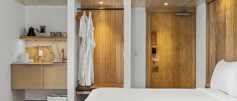 A modern hotel room with a wooden wardrobe, a bed, a small desk with a kettle, and a bench.
