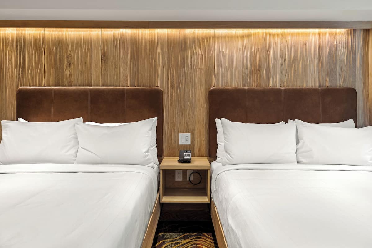 A hotel room with two beds, a wooden headboard, a nightstand with a phone, and a power outlet.