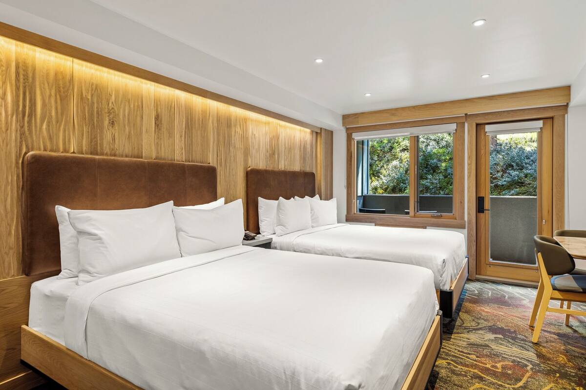 A hotel room with two beds, a wooden headboard, a large window, and a view of greenery.