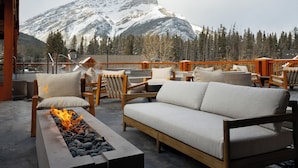 A terrace with a sofa, chairs, and a fire feature, overlooking a snow-covered mountain. - Otter Hotel (Banff)