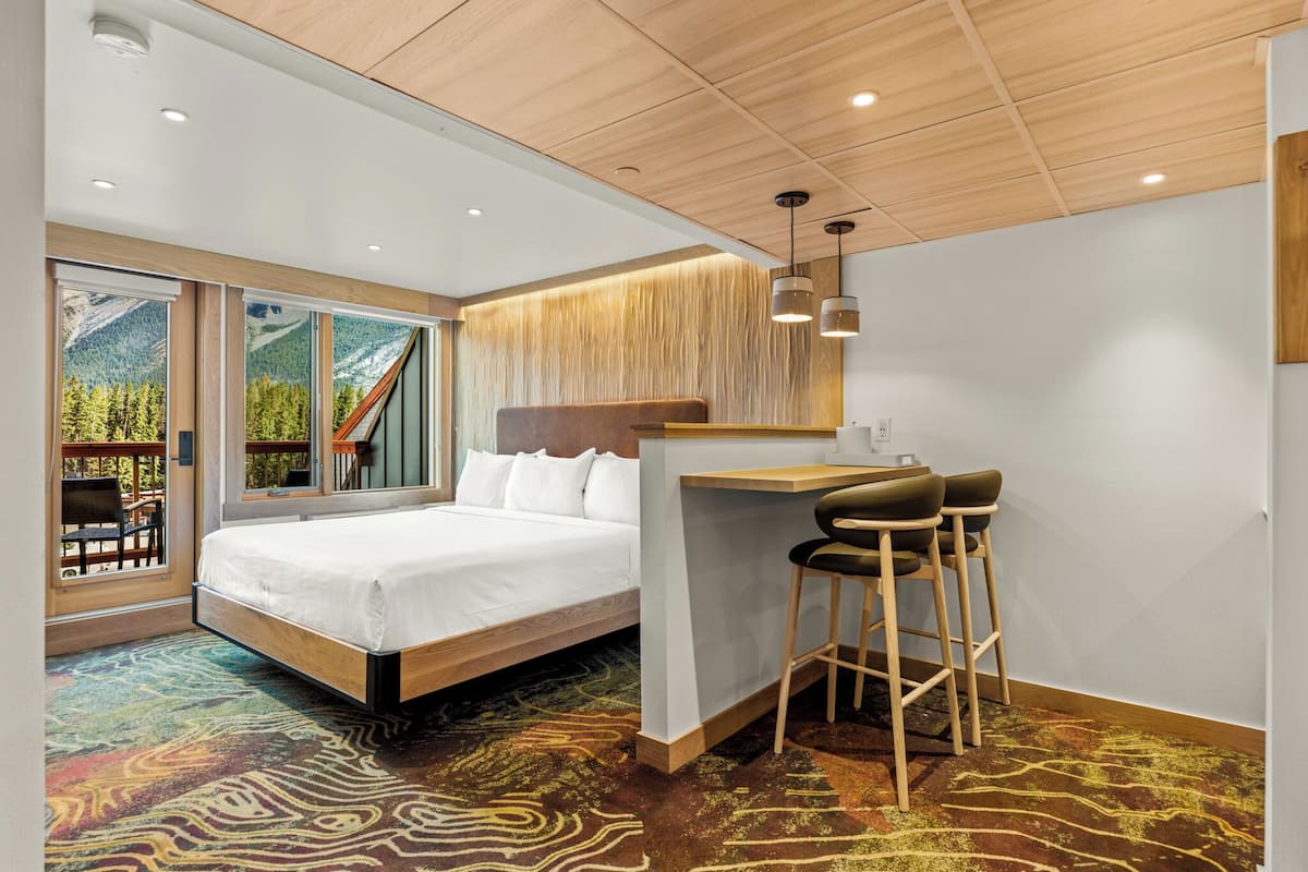 A modern hotel room with a large bed, a desk with two stools, and a view of a mountainous landscape through the windows.