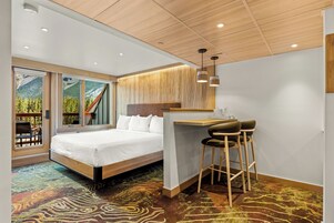 Habitación de hotel moderna con una cama grande, un escritorio con dos taburetes y vistas a un paisaje montañoso por las ventanas.