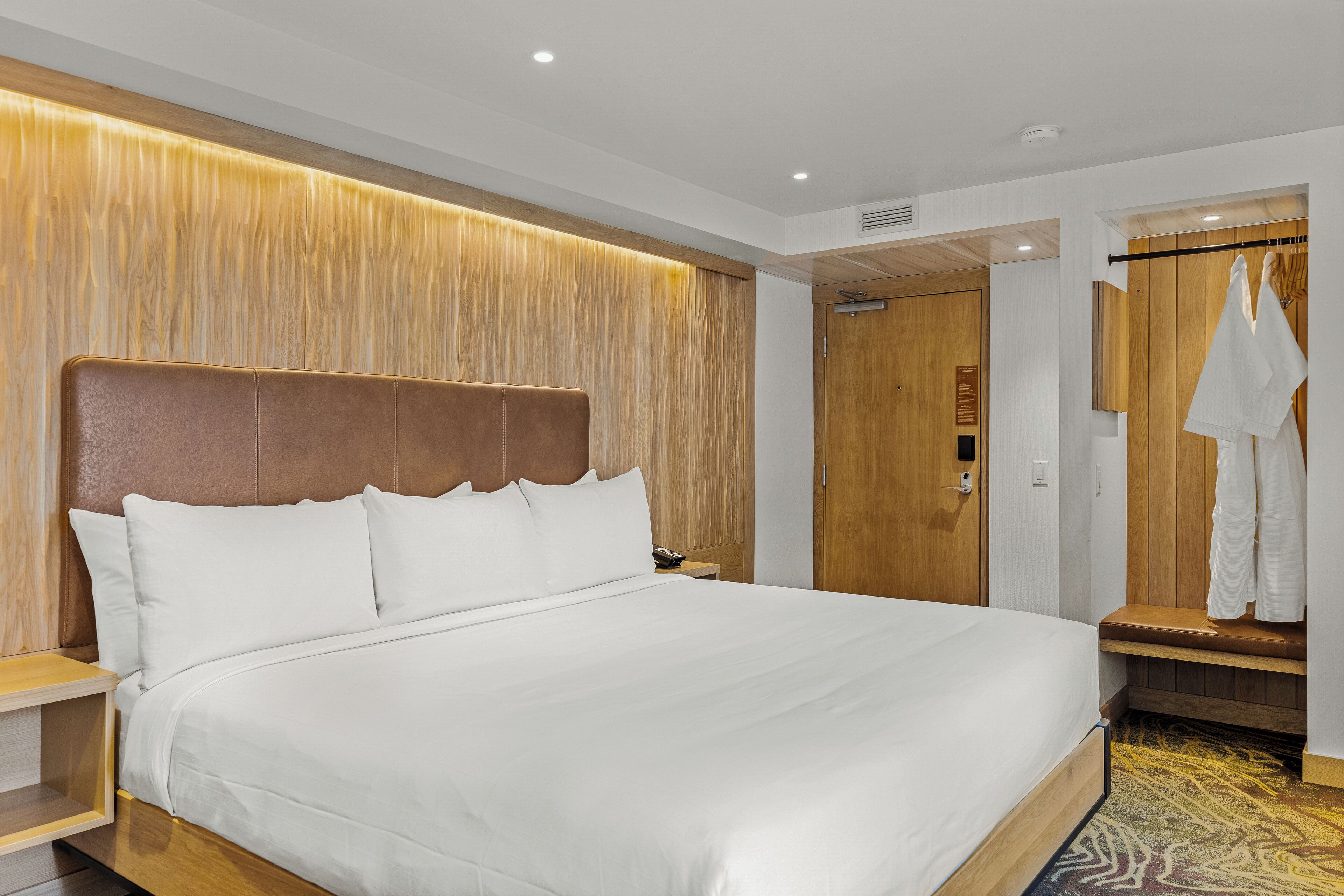 A modern hotel room with a large bed, wooden headboard, and a walk-in closet with a bench.