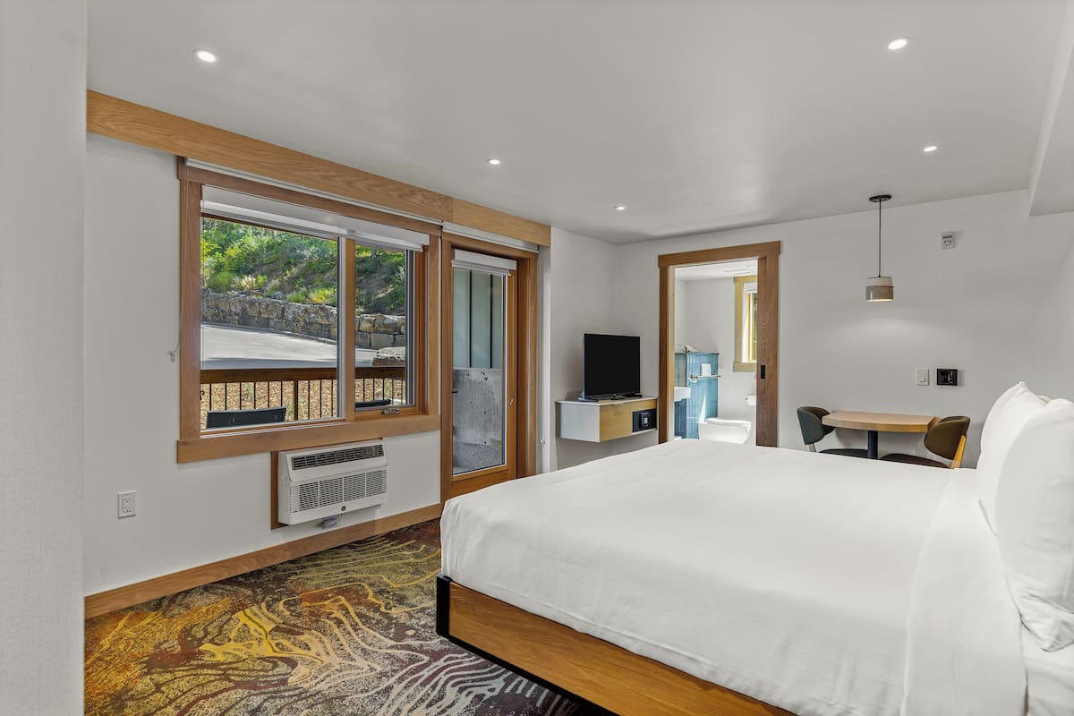 A modern hotel room with a large bed, a desk with a chair, a television, and a window with a view of greenery outside.