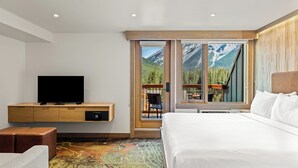 A hotel room with a bed, a TV on a wooden console, a large window with a mountain view, and a colorful carpet. - Otter Hotel (Banff)