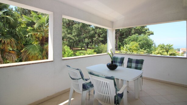 A sunlit patio with a white table and chairs, a vase, and a view of lush greenery and trees. - Charming flat just 350 metres from the beach with private balcony and sea view (Porec)