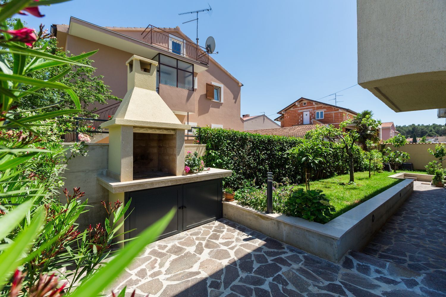 A patio with a stone floor, a barbecue grill, and a garden with various plants and a hedge.