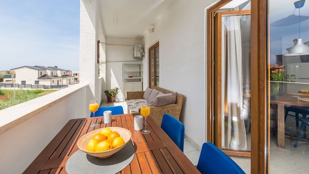 A balcony with a wooden table, blue chairs, a wicker sofa, and a glass of orange juice. - Modern flat with stylish furnishings, sea view, private balcony, barbecue, air conditioning, WiFi (Porec)