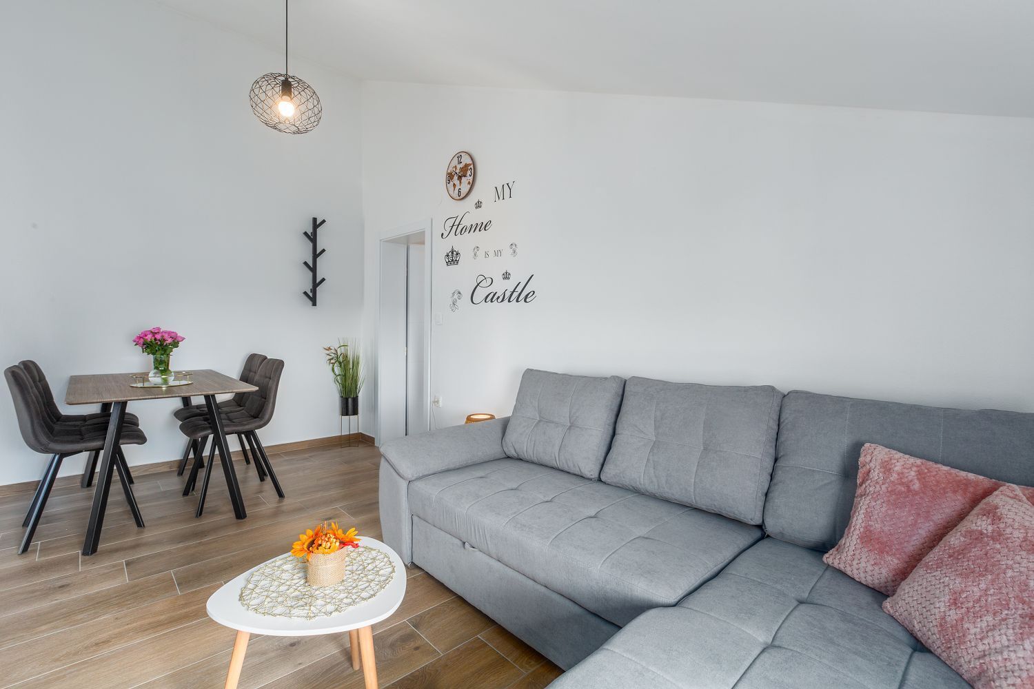 Een moderne woonkamer met een grijze bank, een eettafel met stoelen, een klein rond tafeltje met een bloevas en een wandklok.