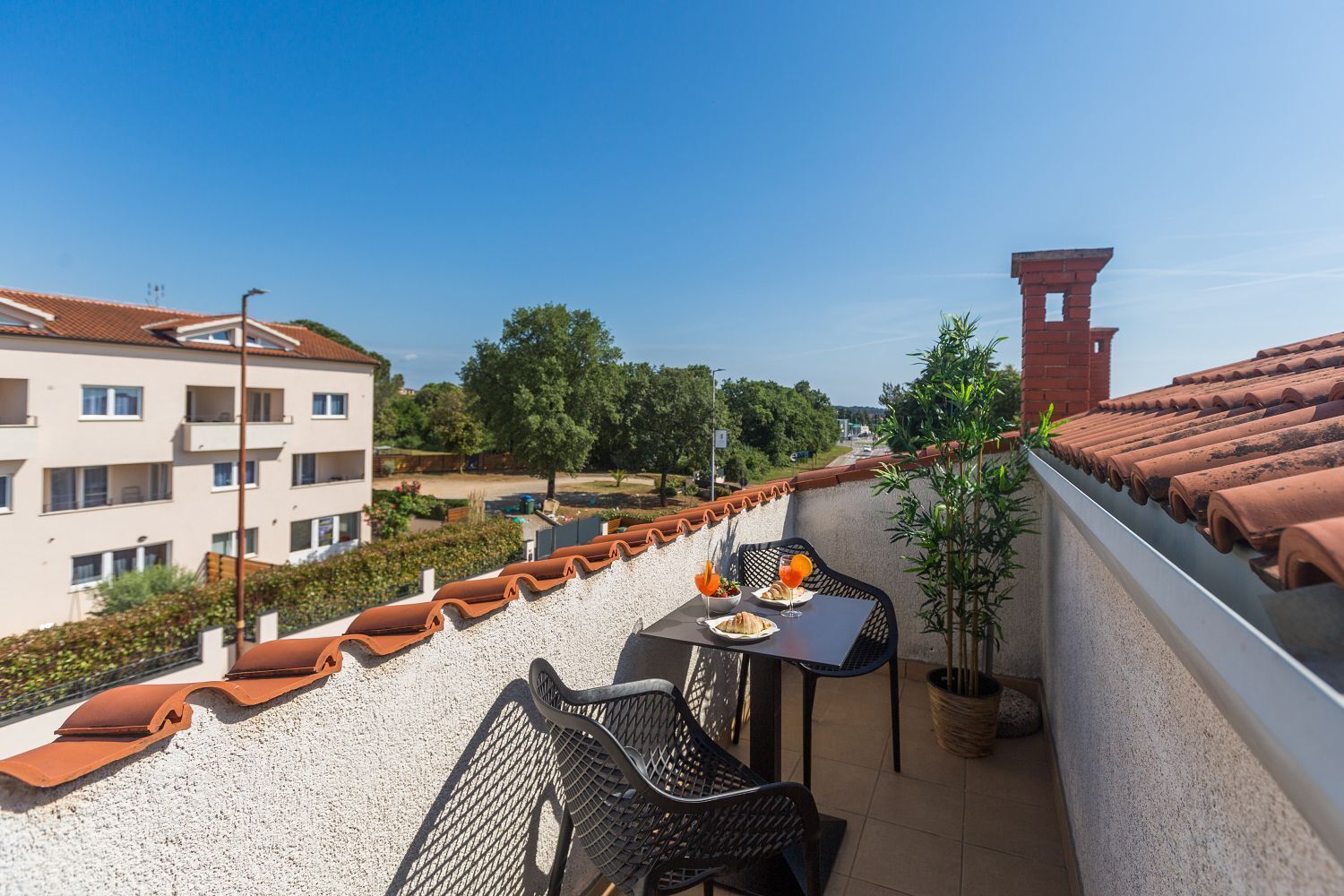A rooftop terrace with a table and chairs, a potted plant, and a view of buildings and trees.