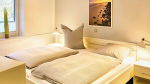 Ein Schlafzimmer mit zwei Betten, einem Fenster mit Jalousien, einer Nachttischlampe und einem gerahmten Bild an der Wand.