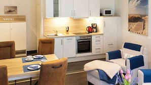 Eine moderne Küche mit weißen Schränken, einer Mikrowelle und einer Kaffeemaschine. Es gibt einen Esstisch mit Stühlen und ein Sofa mit einer Decke.