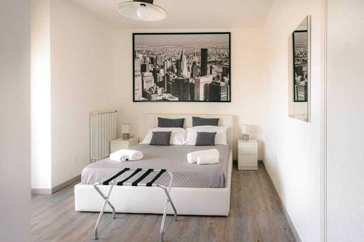 Une chambre moderne avec un grand lit, deux tables de chevet, un radiateur et un tableau représentant un paysage urbain au-dessus du lit.