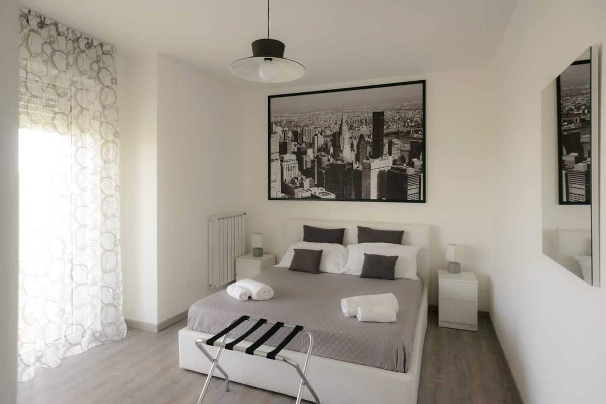 Une chambre d’hôtel moderne avec un grand lit, deux tables de chevet, un radiateur et une œuvre d’art représentant un paysage urbain accrochée au mur.