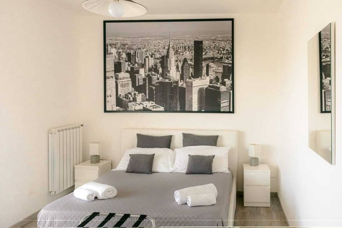 Une chambre moderne avec un grand lit, une œuvre d’art représentant un paysage urbain, un luminaire suspendu et un plancher en bois.