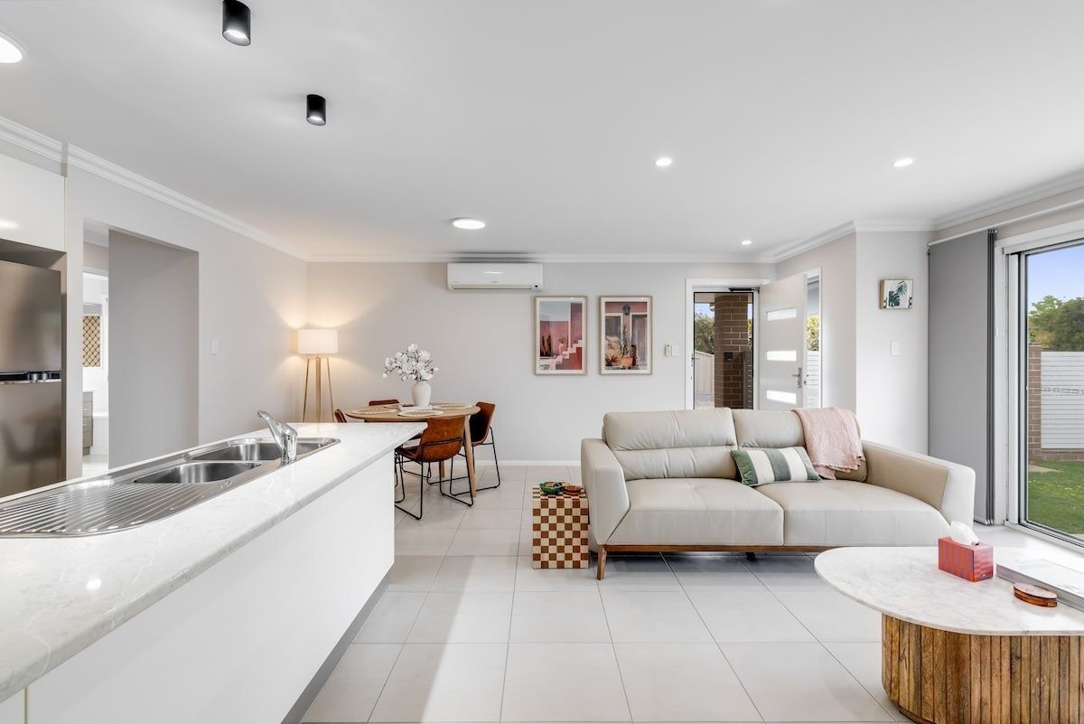Una cocina moderna con un lavamanos integrado, una zona de comedor con mesa y sillas, un sofá color beige y un ventanal con vistas al exterior.