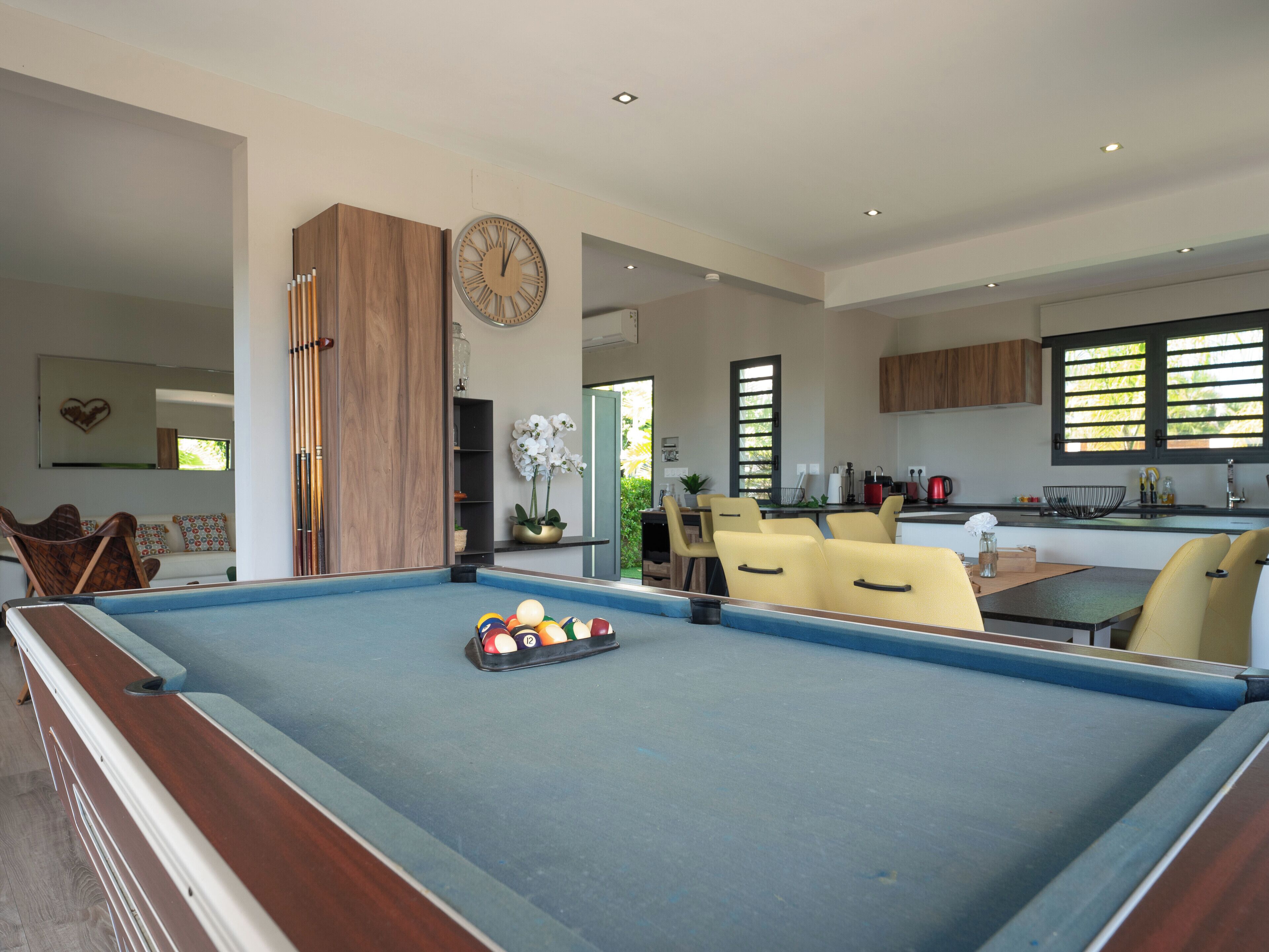 A modern living room with a pool table, yellow chairs, a dining area, and a kitchen.
