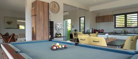 A modern living room with a pool table, yellow chairs, a dining area, and a kitchen.