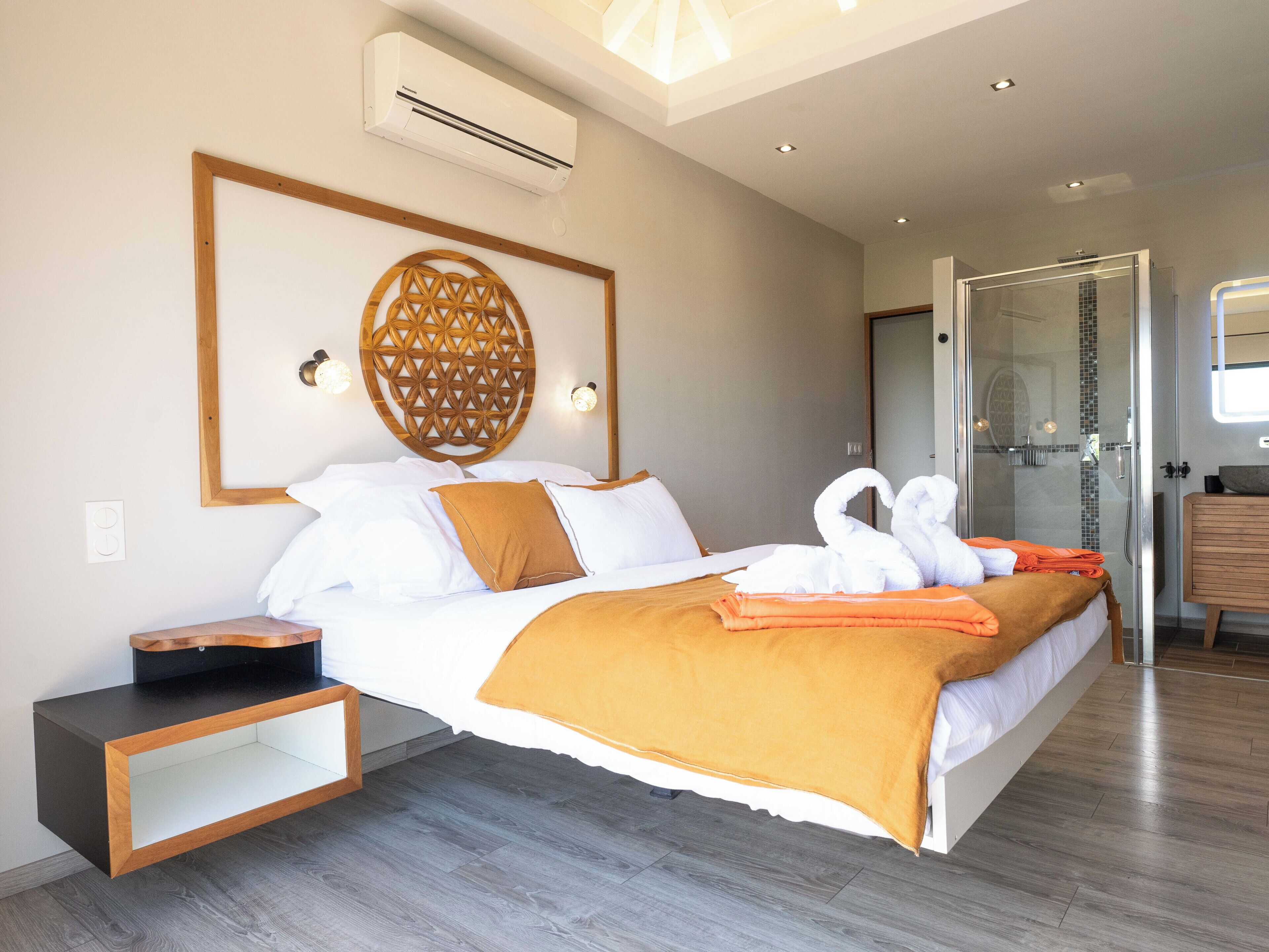Une chambre d’hôtel moderne dotée d’un grand lit, d’une tête de lit en bois, d’un climatiseur, d’une table de chevet et d’une salle de bain avec une douche en verre.