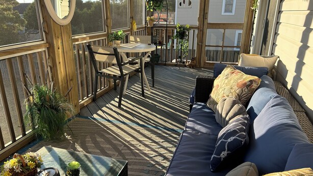 Eine sonnendurchflutete Terrassenduschel mit einem Holztisch, Stühlen und einem blauen Sofa mit Kissen.