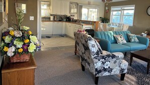Ein geräumiges Wohnzimmer mit einem petrolfarbenen Sofa, einem gemusterten Sessel und einem Couchtisch aus Holz. Im Hintergrund ist eine Küche mit weißen Schränken und ein Essbereich mit Tisch und Stühlen zu sehen.