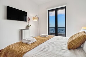 A modern hotel room with a large bed, a flat-screen TV mounted on the wall, a bedside lamp, and a view of the sea through a sliding door.