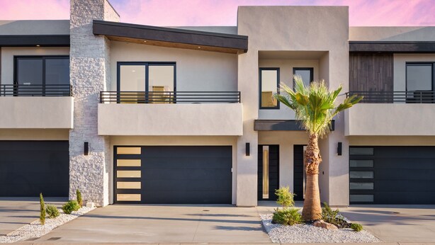 A modern two-story house with a garage, a balcony, and a small palm tree in the front yard.