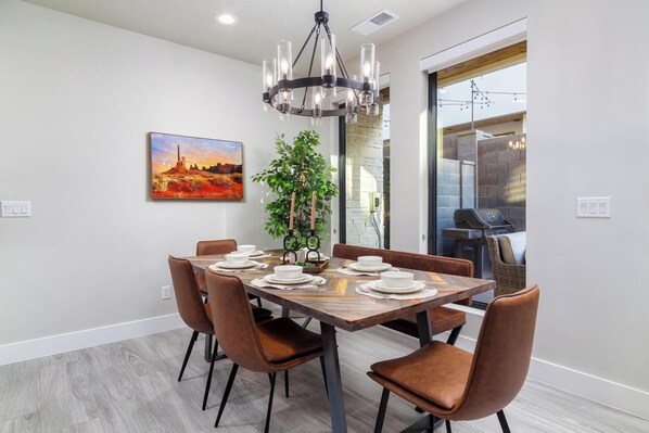 A modern dining area with a wooden table set for six, surrounded by brown chairs. A chandelier hangs above, and a painting of a sunset is on the wall.