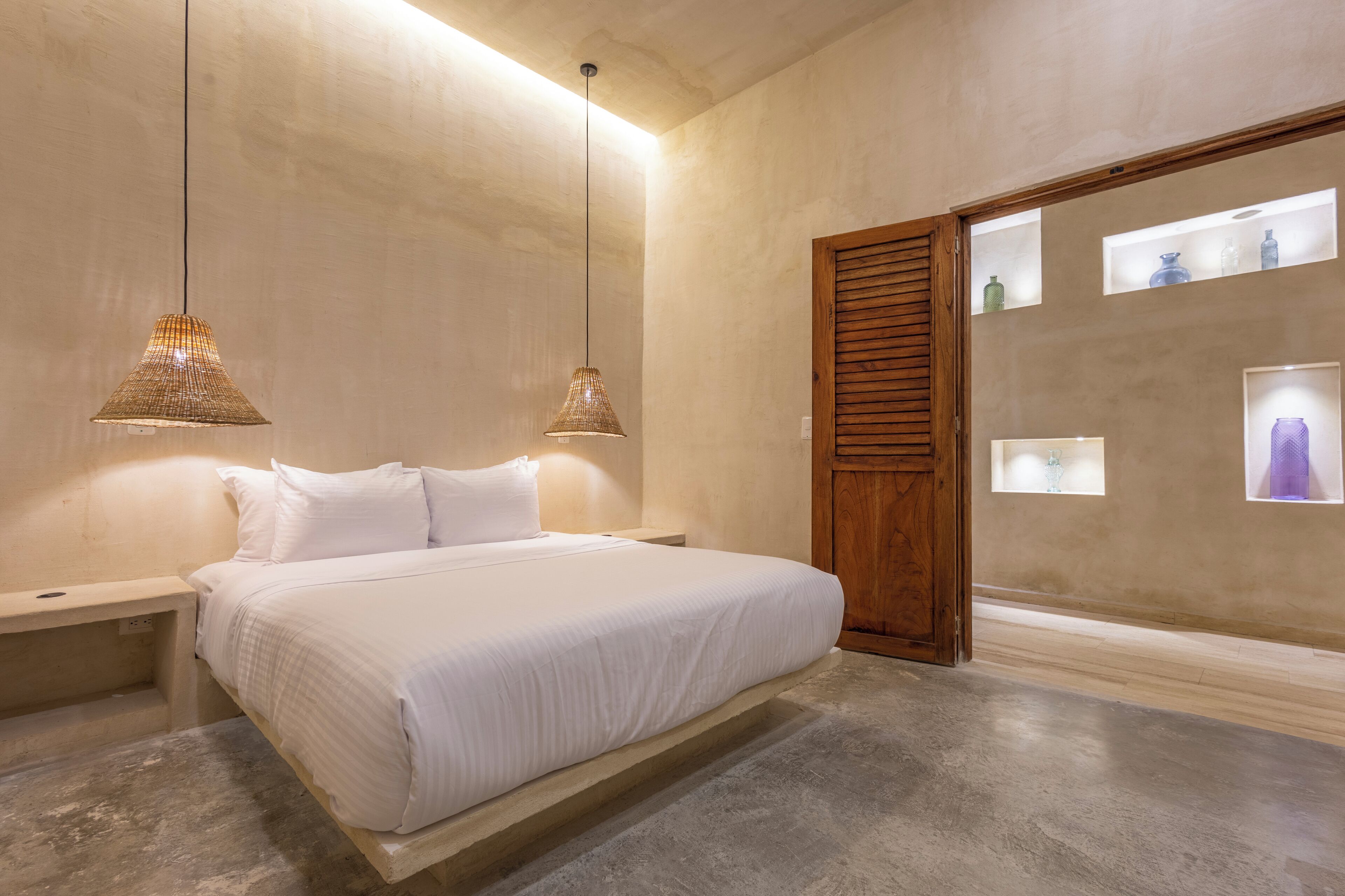 Un dormitorio con cama, mesitas de noche, lámparas colgantes, una puerta de madera y estantes en la pared con objetos decorativos.