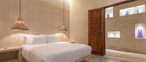 Un dormitorio con cama, mesitas de noche, lámparas colgantes, una puerta de madera y estantes en la pared con objetos decorativos.