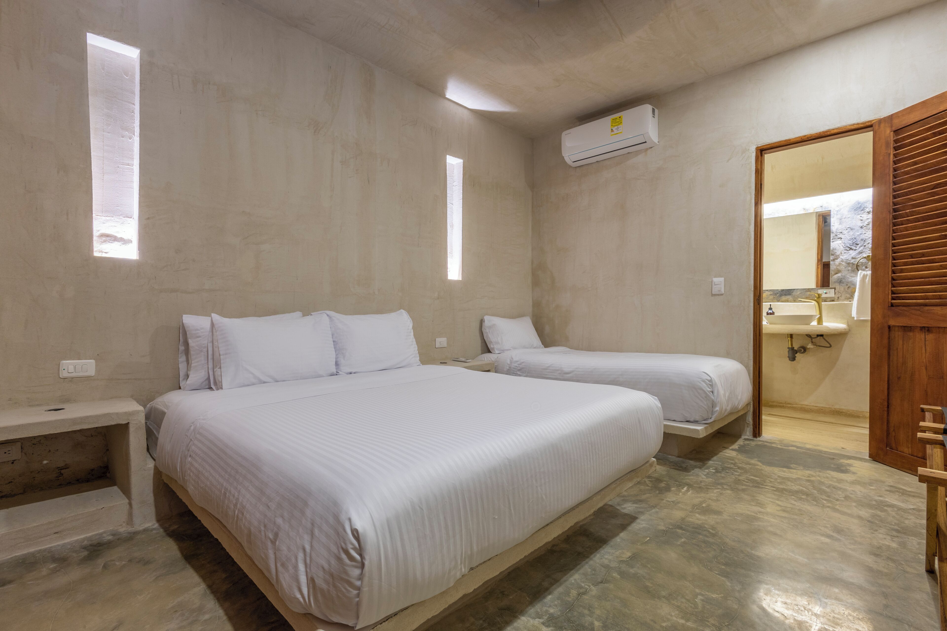 A bedroom with two beds, a wooden bench, a small table, a window, and a bathroom with a sink and mirror.