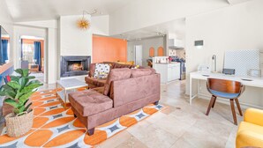 Een moderne woonkamer met een bank, een salontafel en een open haard. Achtergrond toont een keuken met een eettafel en stoelen.