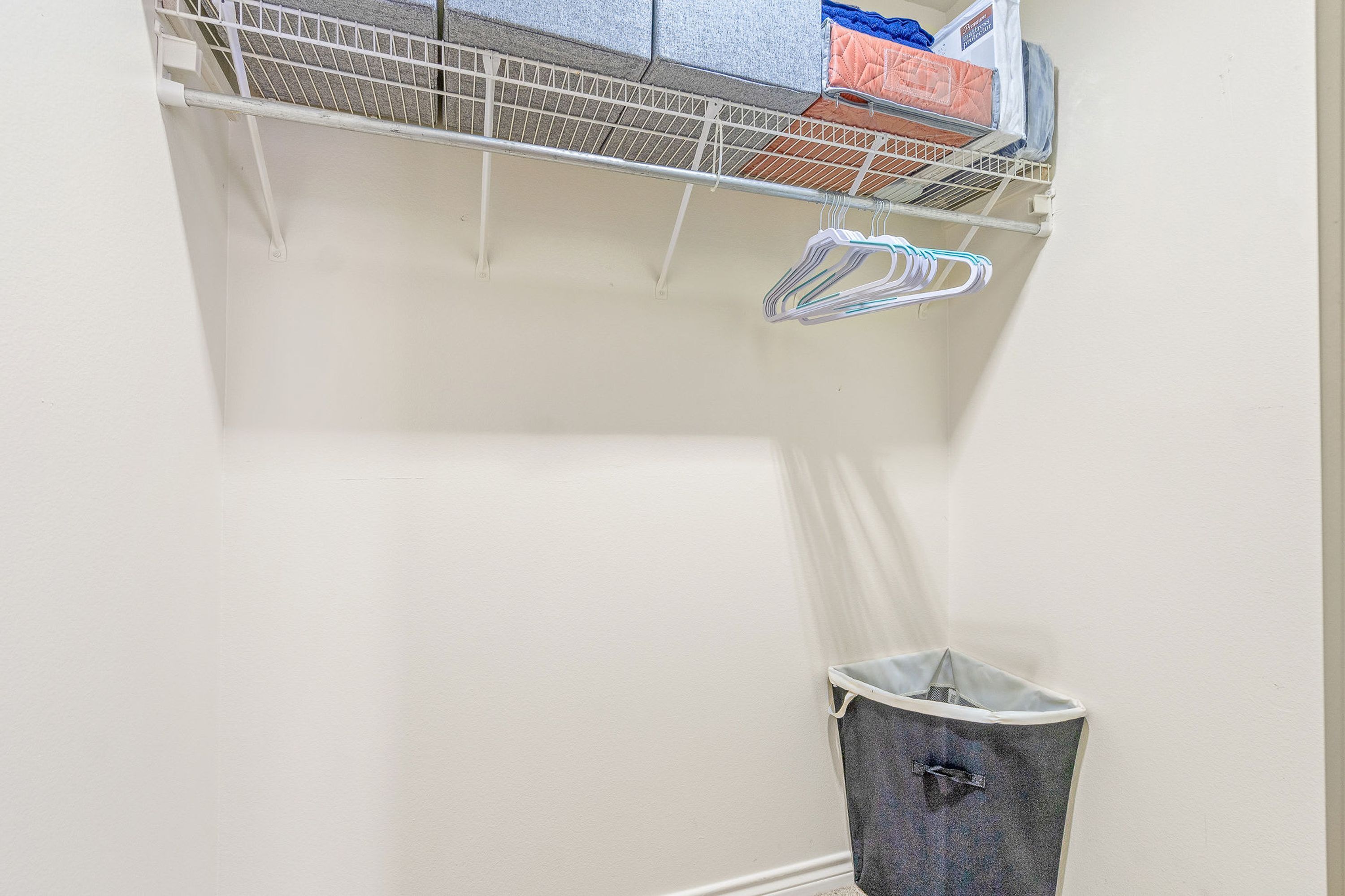 A laundry area with a metal rack, a grey trash bin, and a wall-mounted shelf with folded laundry.