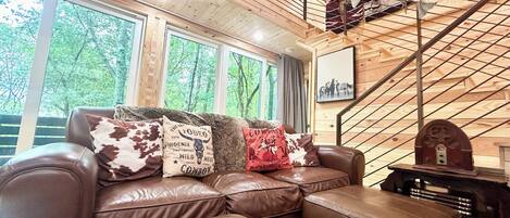 A cozy living room with a leather sofa, a staircase, and a view of trees through large windows.
