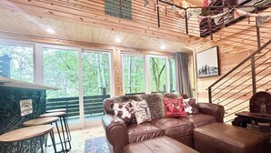 Ein gemütliches Wohnzimmer mit einem braunen Ledersofa, einem Holztisch und einer Bar mit Hockern. Große Fenster bieten einen Blick ins Freie.