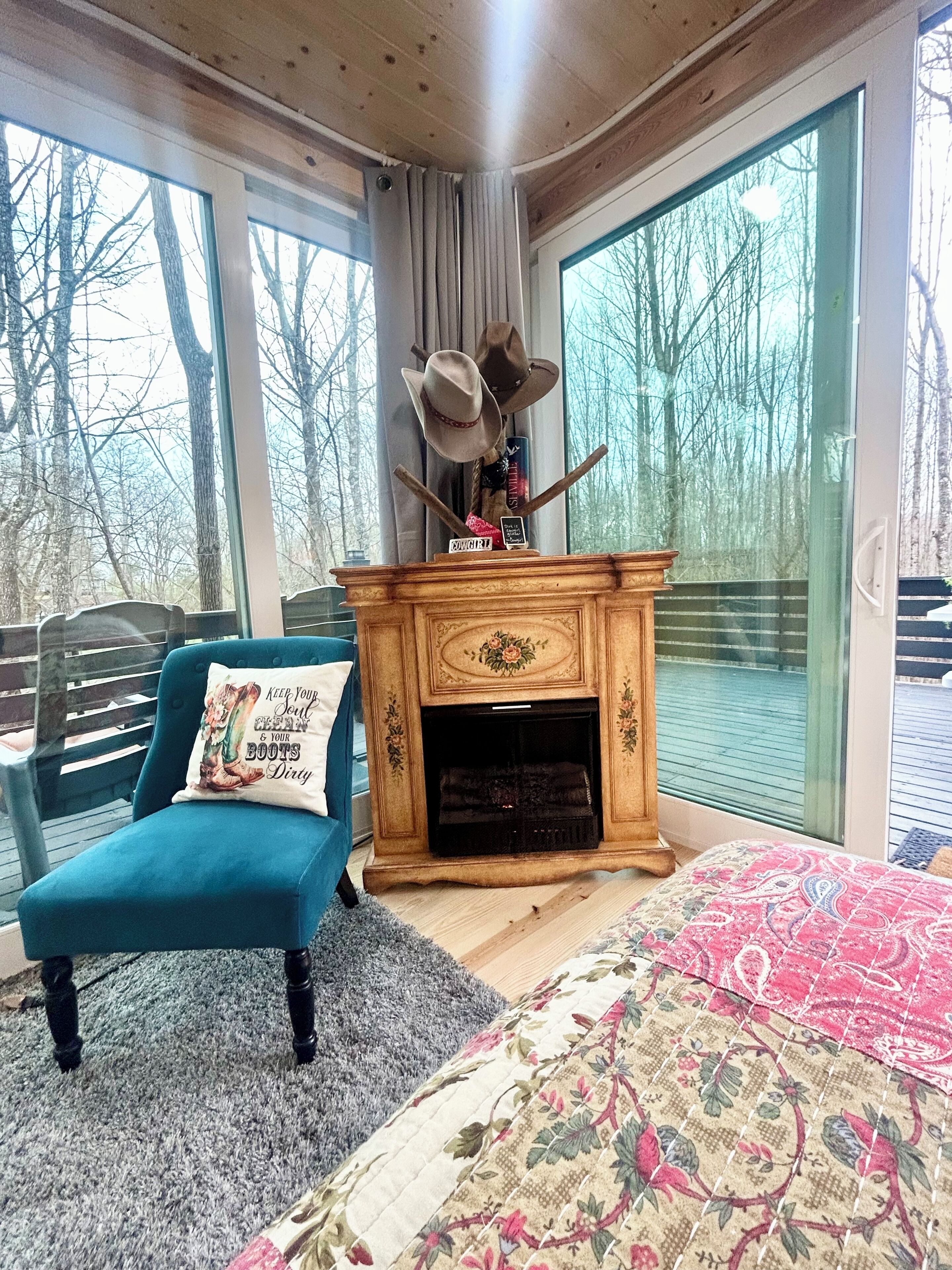 A room with a fireplace, a blue armchair, a bed, and large windows offering a view of trees.
