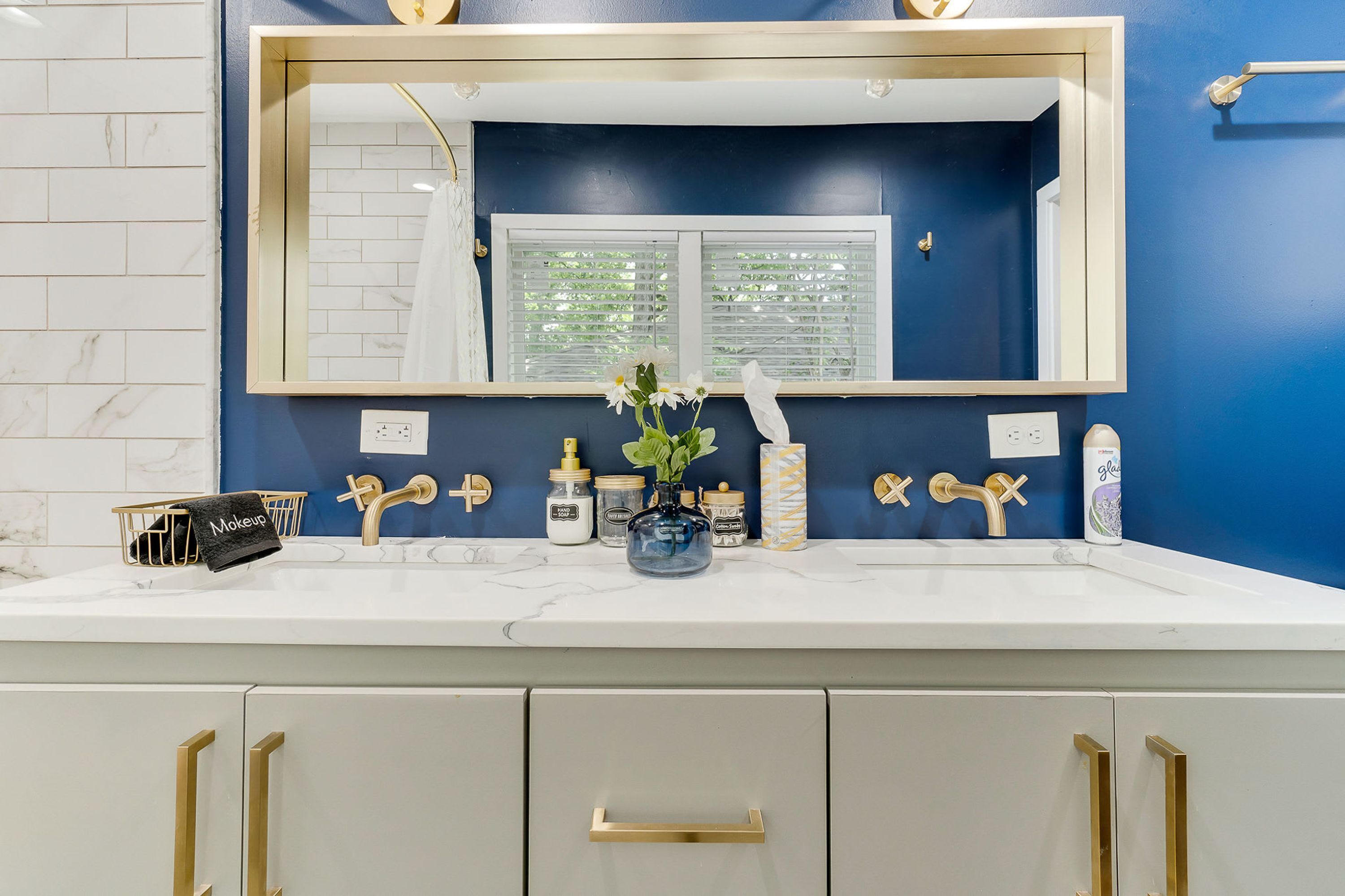 A bathroom with a white marble countertop, gold handles, a large mirror, and a window with blinds.