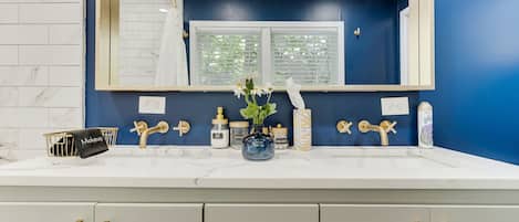 A bathroom with a white marble countertop, gold handles, a large mirror, and a window with blinds.