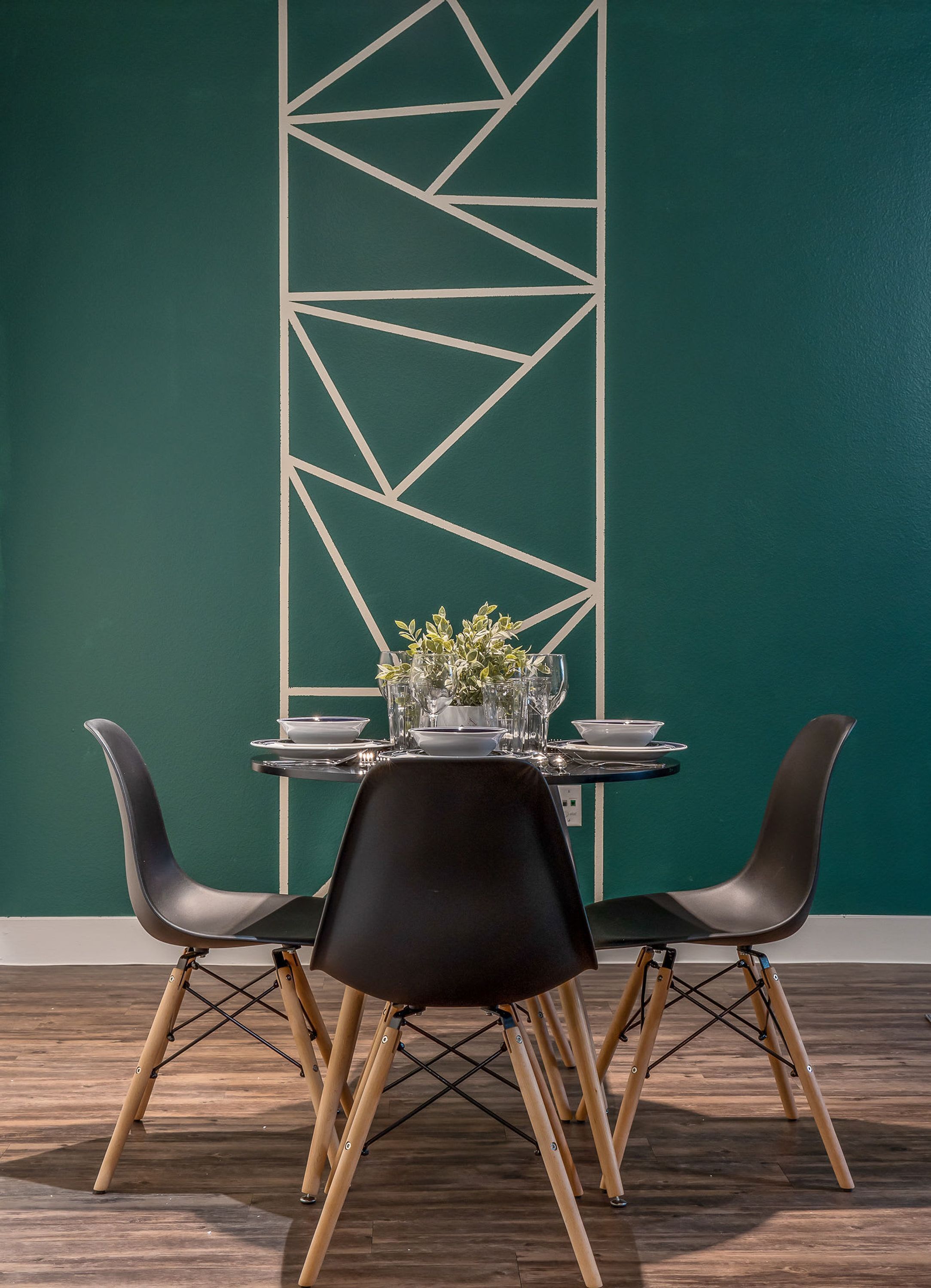 Un comedor con una mesa dispuesta para dos personas, dos sillas negras con patas de madera, y una pared decorativa con diseños geométricos.