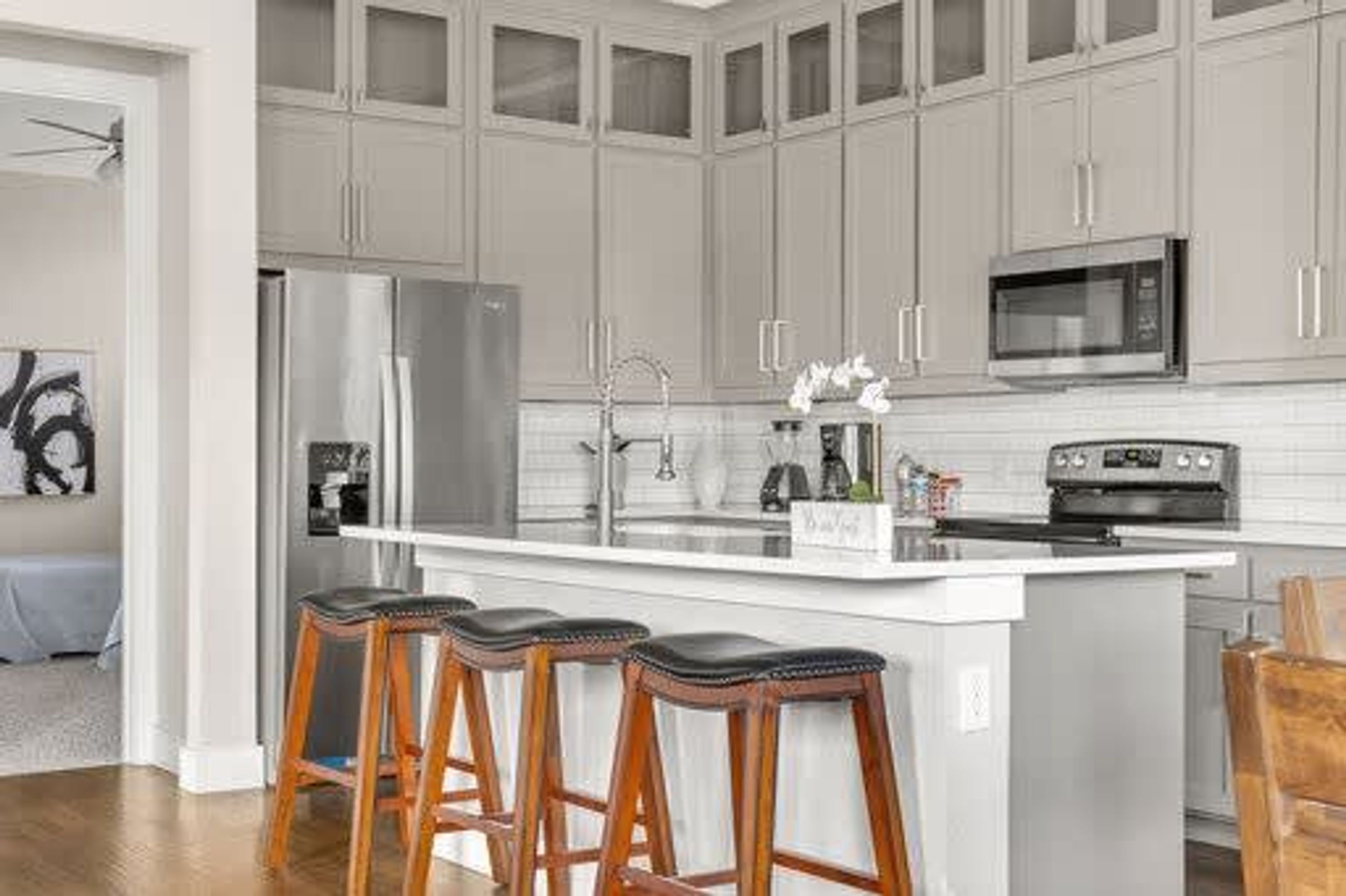 A kitchen with grey cabinets, a stainless steel refrigerator, a microwave, a sink, and a bar stool area.