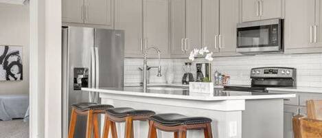 A kitchen with grey cabinets, a stainless steel refrigerator, a microwave, a sink, and a bar stool area.