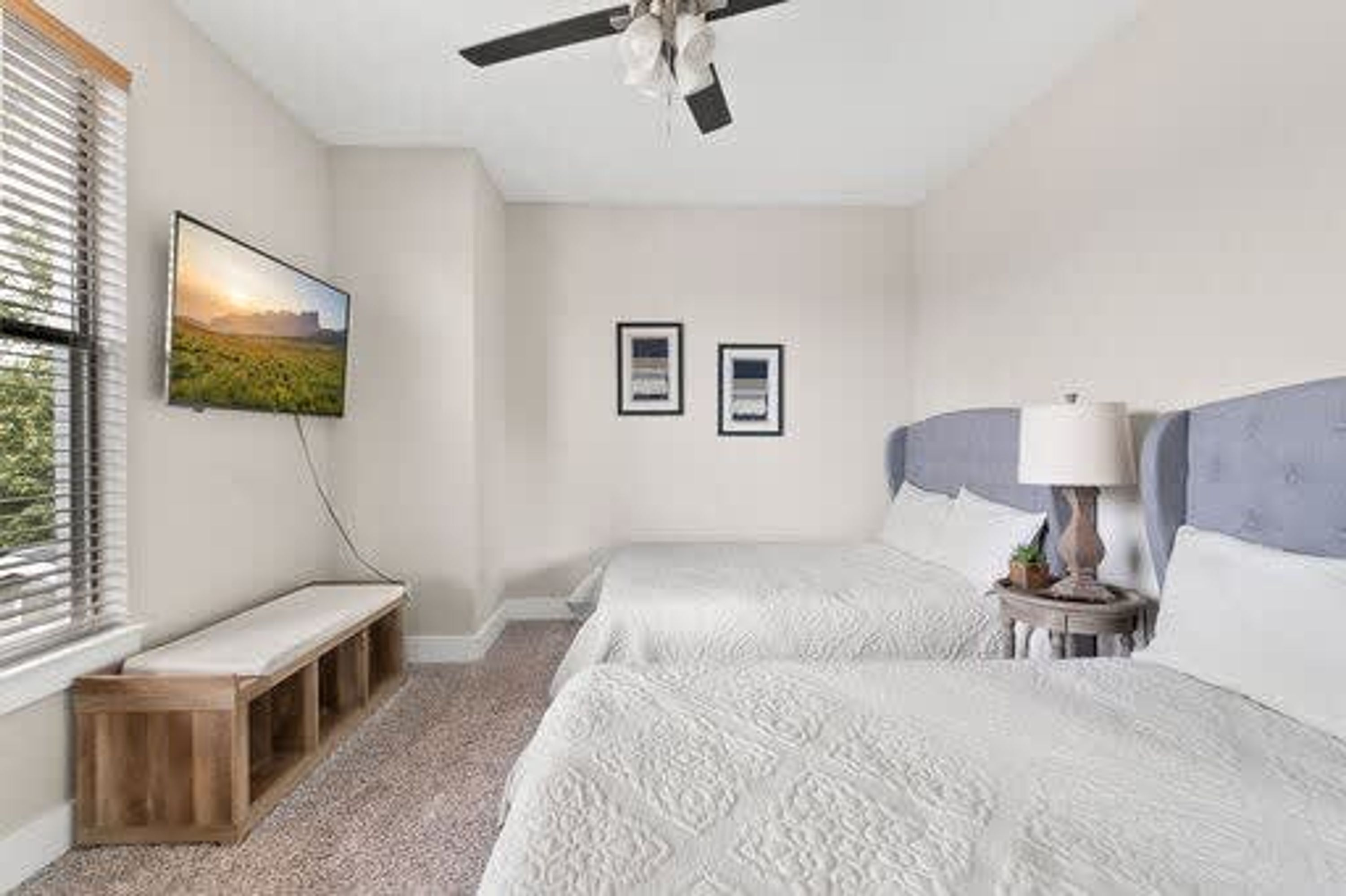 A bedroom with two beds, a ceiling fan, a bench, a TV, and framed pictures on the wall.