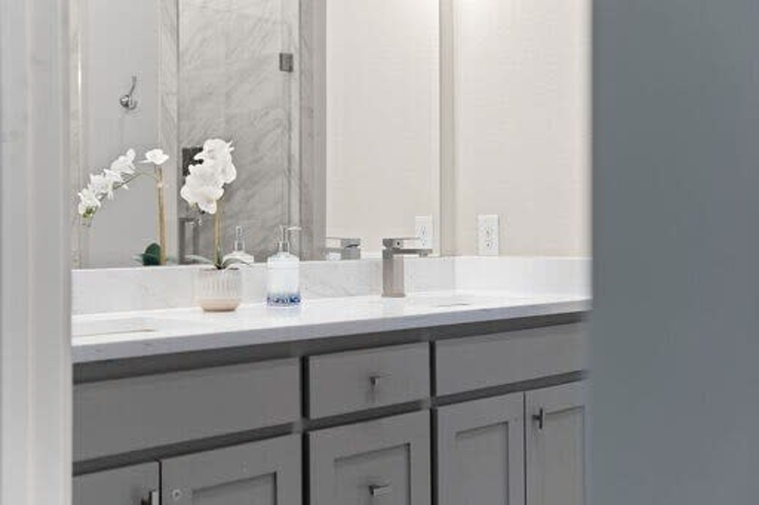 A bathroom with a large mirror, white sink, and grey cabinets.