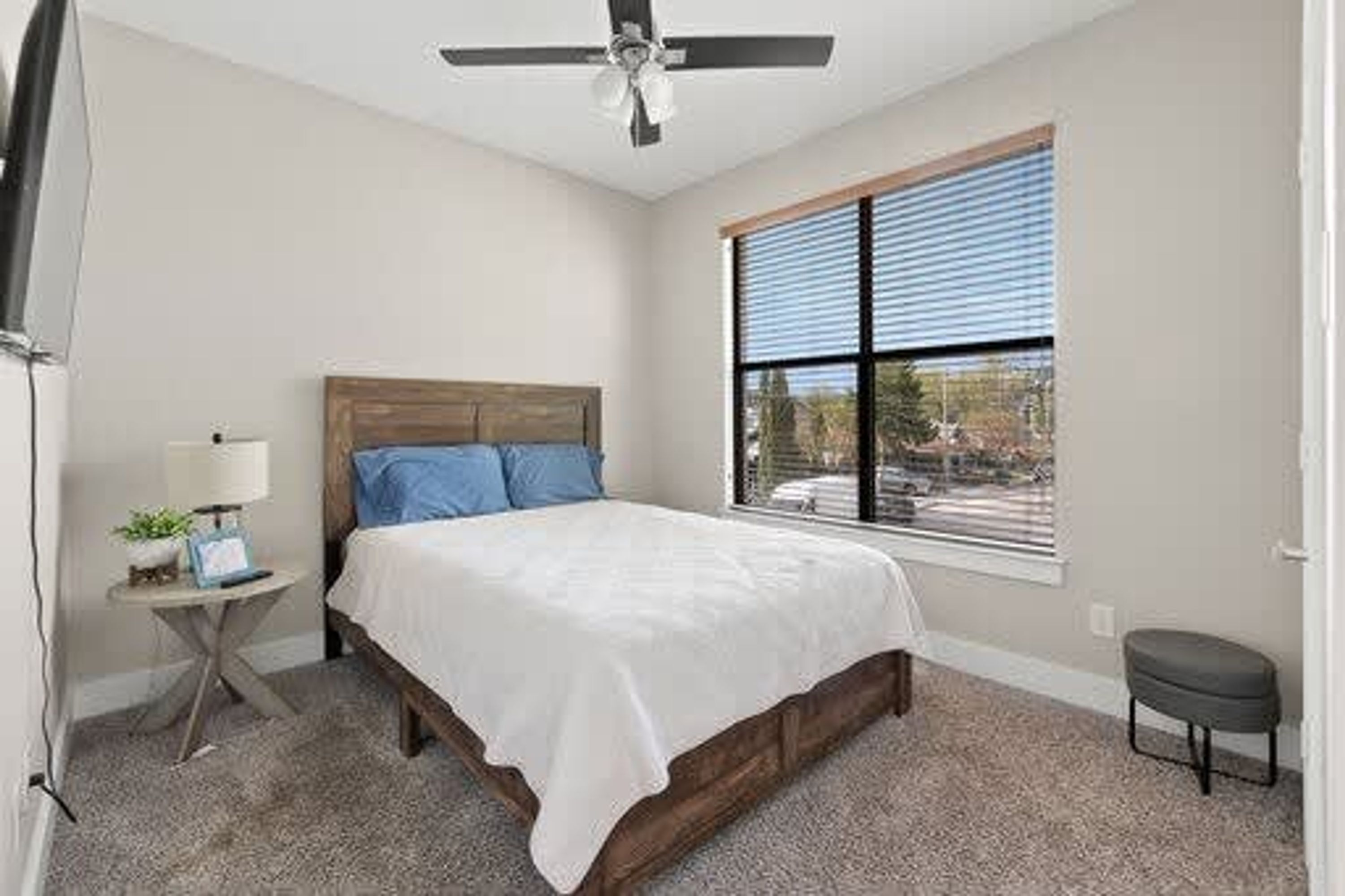 A bedroom with a bed, a ceiling fan, a window with blinds, a small table with a lamp, and a bench.