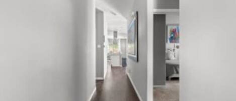 A hallway with a wooden floor and white walls, leading to a room with a bed and a painting on the wall.