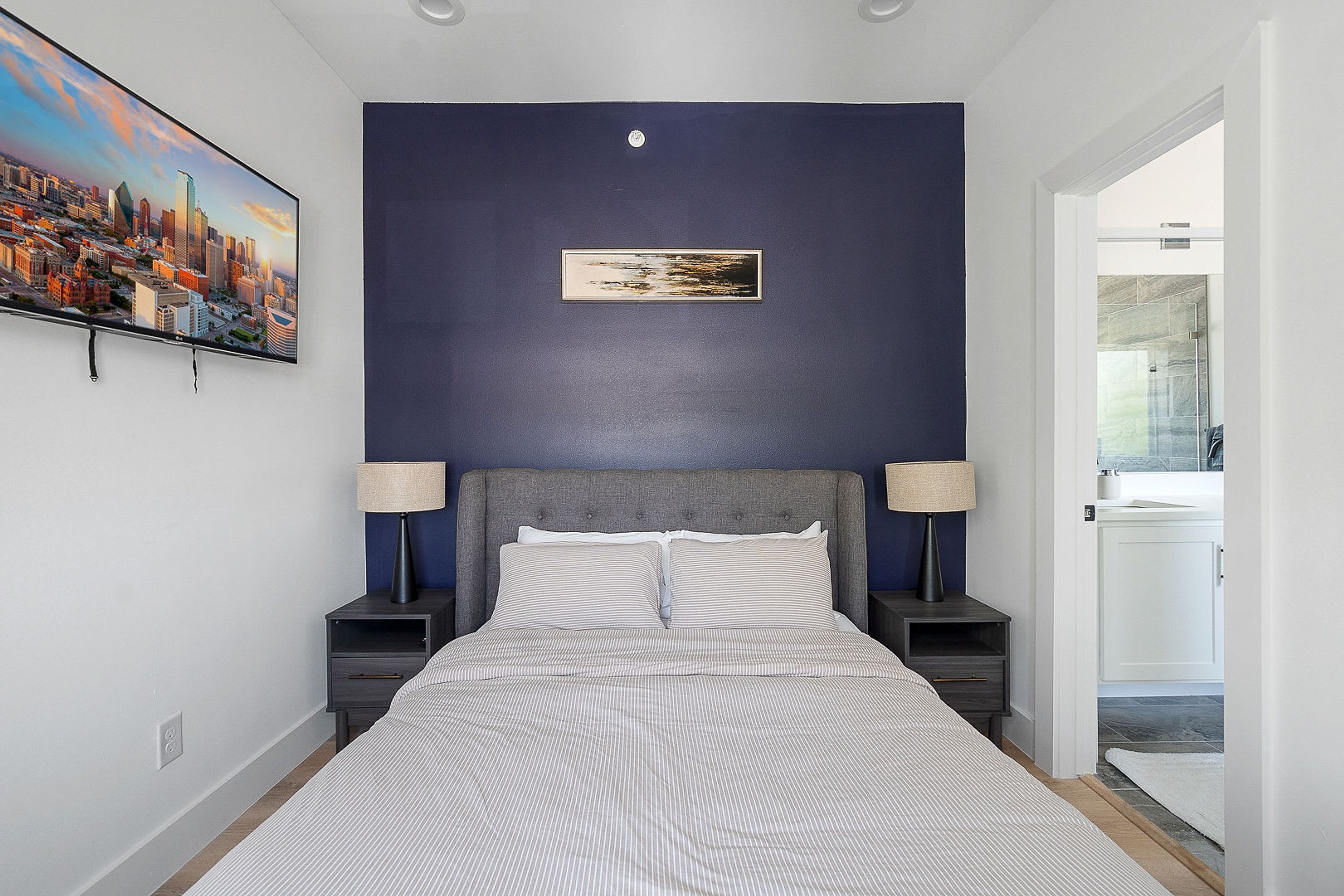 A bedroom with a cityscape painting, a headboard, two bedside tables with lamps, and a view of the bathroom.