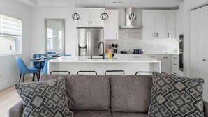 A modern kitchen with white cabinets, a central island, and pendant lighting. There is a dining area with blue chairs and a sofa with patterned cushions.