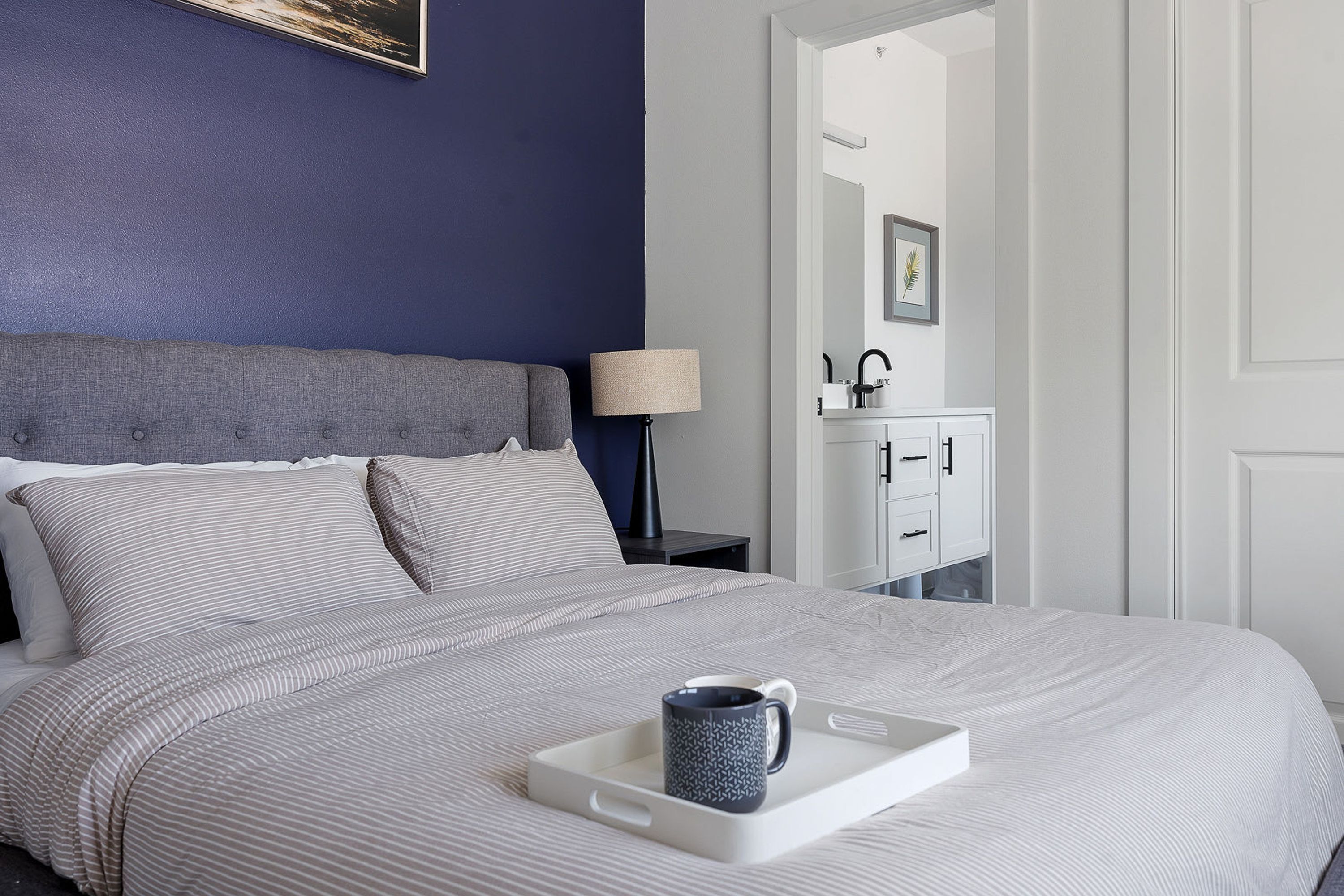 Un lit bien fait, avec un plateau sur lequel se trouvent une tasse, une lampe de chevet et une commode dotée d’un miroir.