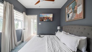 A bedroom with a bed, a ceiling fan, a cityscape painting, and a window with curtains. - Urban Comfort King Bed Rooftop Backyard (Dallas)